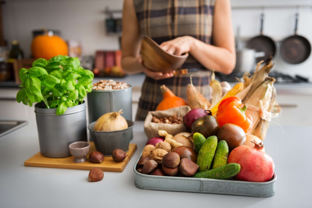 Table de cuisine remplit de légumes, herbes et noix. Une femme en arrière plan se prépare à cuisiner ces ingrédients.